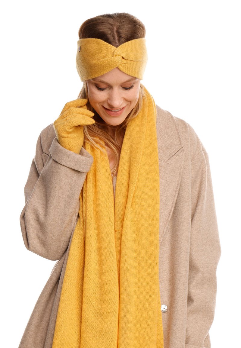 Gul stickad hårband med en tvistad design, matchande handskar och en lång scarf. Mjuk textur, bärs med en beige kappa.
