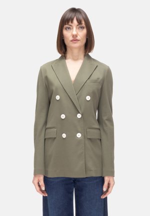 Donna con capelli castano lisci indossa un blazer doppiopetto verde oliva con sei bottoni bianchi e jeans blu, in piedi davanti a uno sfondo bianco.