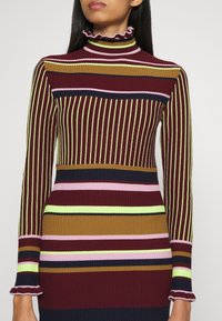 Ribbad långärmad polotröja i bordeaux med flerfärgade vertikala ränder i grönt, brunt, rosa och marinblått. Volangkrage och volang ärmar.