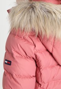 Doudoune rose clair avec un col en fausse fourrure beige duveteuse et des coutures visibles. Présente un écusson avec un logo en bleu marine, rouge et blanc sur le côté.