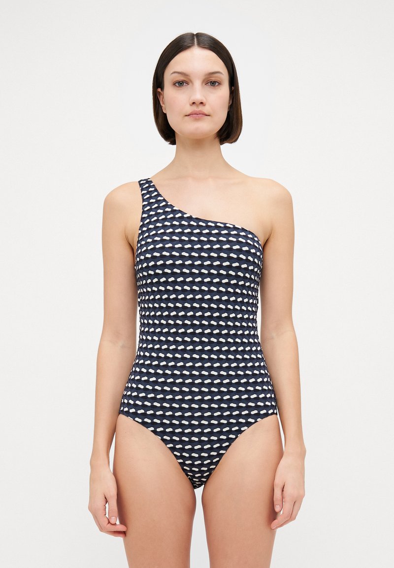 Femme portant un maillot de bain à une épaule, bleu marine et blanc à motifs, se tenant contre un fond blanc uni.