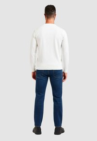 Camicia bianca a maniche lunghe con orlo a coste, abbinata a jeans blu e scarpe nere. Vista da dietro, che mette in risalto il design pulito della camicia.