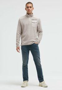 Helle graue Pullover mit halbem Reißverschluss-Kragen und Brusttasche, kombiniert mit blauen Jeans und weißen Turnschuhen, zeichnet sich durch strukturierten Stoff und dezente Markenkennzeichnung aus.