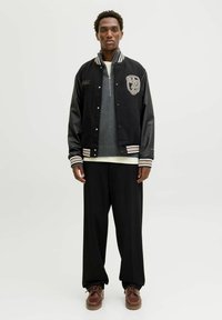 Ung mand iført en sort varsity-jacke med læderærmer, grå zip-sweater, løse sorte bukser og brune snøresko på en hvid baggrund.