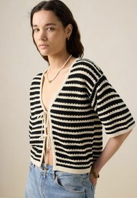 Cardigan tricoté noir et crème à rayures avec des manches courtes, doté d'un décolleté en V profond et d'une fermeture à nouer sur le devant, associé à un jean bleu clair.