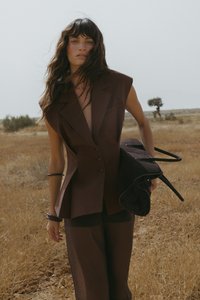 Brūns pielāgots veste ar plašiem pleciem, vienkrūtējo pogu aizdare, apvienota ar brīviem brūniem biksēm, tur melnu somu ar rokturiem.