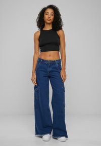 Zwarte cropped tanktop en wijdvallende blauwe denim cargobroek met meerdere zakken en gefranjerde zoom. Model draagt witte sneakers.
