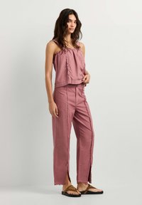 Woman stands wearing sleeveless pink top and matching pants with front slits and frayed edges, paired with brown sandals.