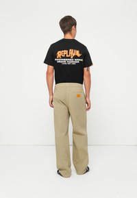 Man facing away wearing black graphic t-shirt with orange text and beige loose pants with orange tag, standing on white background.