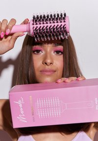 Cepillo de pelo redondo rosa con cerdas negras, sostenido sobre la cabeza de un modelo. La caja del producto es rosa con ilustraciones y texto en blanco.