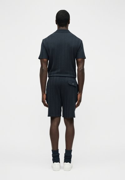 Homme debout de dos, portant une chemise à manches courtes texturée sombre et un short assorti avec une poche arrière boutonnée, des chaussures blanches et des chaussettes foncées.