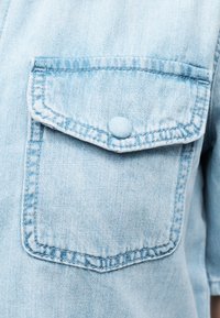 Τσέπη από ανοιχτό μπλε τζιν ύφασμα με καπάκι και κουμπί καλυμμένο με ύφασμα στο ίδιο χρώμα σε ένδυμα.