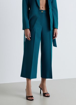 Donna che indossa pantaloni larghi cropped color teal e blazer coordinato, abbinati a tacchi alti neri open-toe, in piedi davanti a uno sfondo semplice.