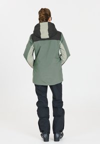 Waterdichte jas in groen en zwart, met een hoge kraag, een capuchon en lange mouwen. Gepaard met zwarte sneeuwbroeken en laarzen.