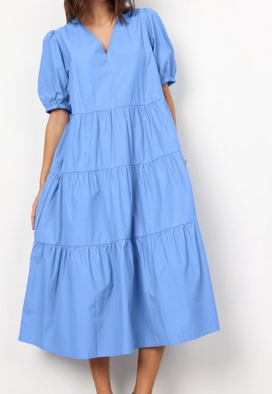 Kvinde iført en løs, lagdelt, lyseblå midi-kjole med puffede korte ærmer og V-udskæring, stående mod en ensfarvet hvid baggrund.