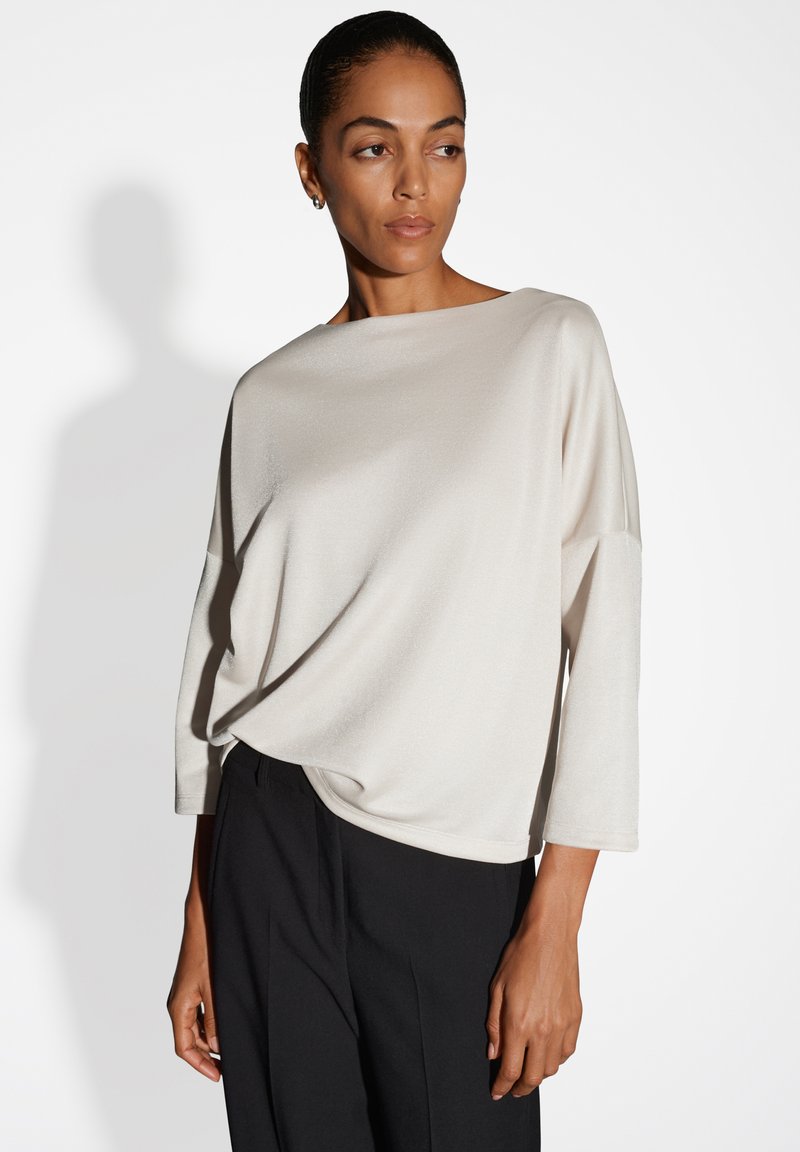 Donna che indossa una maglia lunga e larga di colore avorio con scollatura a barca e pantaloni neri, in posa davanti a uno sfondo bianco semplice.