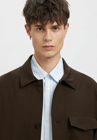 Brun jakke med knappelukking og brystlomme over en lyseblå stripete skjorte. Glatt stoff, strukturert krage og skreddersydd passform.