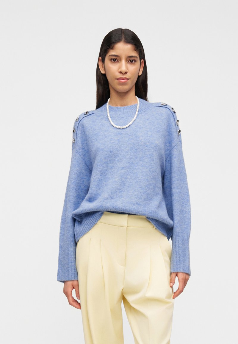 Γυναίκα με μακριά σκούρα μαλλιά φοράει ανοιχτό μπλε πουλόβερ με κουμπιά στον ώμο, κρεμ πλισέ παντελόνι και μονόσκοινο μαργαριταρένιο κολιέ, όρθια.