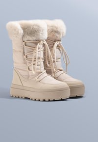 Beige winterlaarzen met nepbont, gewatteerde schacht, veterluiting aan de voorkant en dikke rubberen zool met een textuurpatroon voor grip.