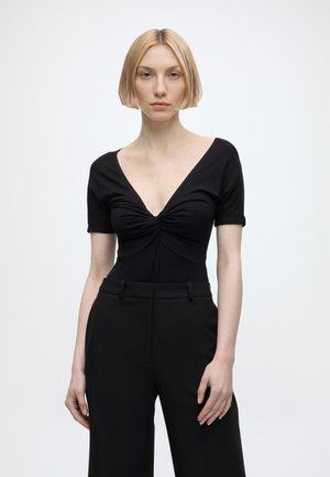 Femme aux cheveux blonds courts porte un haut noir décolleté en V profond avec fronces et un pantalon noir taille haute, debout devant un fond uni.