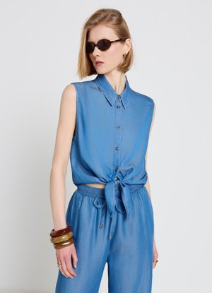 Femme portant une chemise bleue sans manches nouée, un pantalon à cordon assorti, des lunettes de soleil foncées et des bracelets épais au poignet gauche.