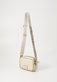 Borsa a tracolla beige realizzata in materiale liscio, con piping nero a contrasto, una cerniera superiore e una tracolla regolabile con branding impresso.