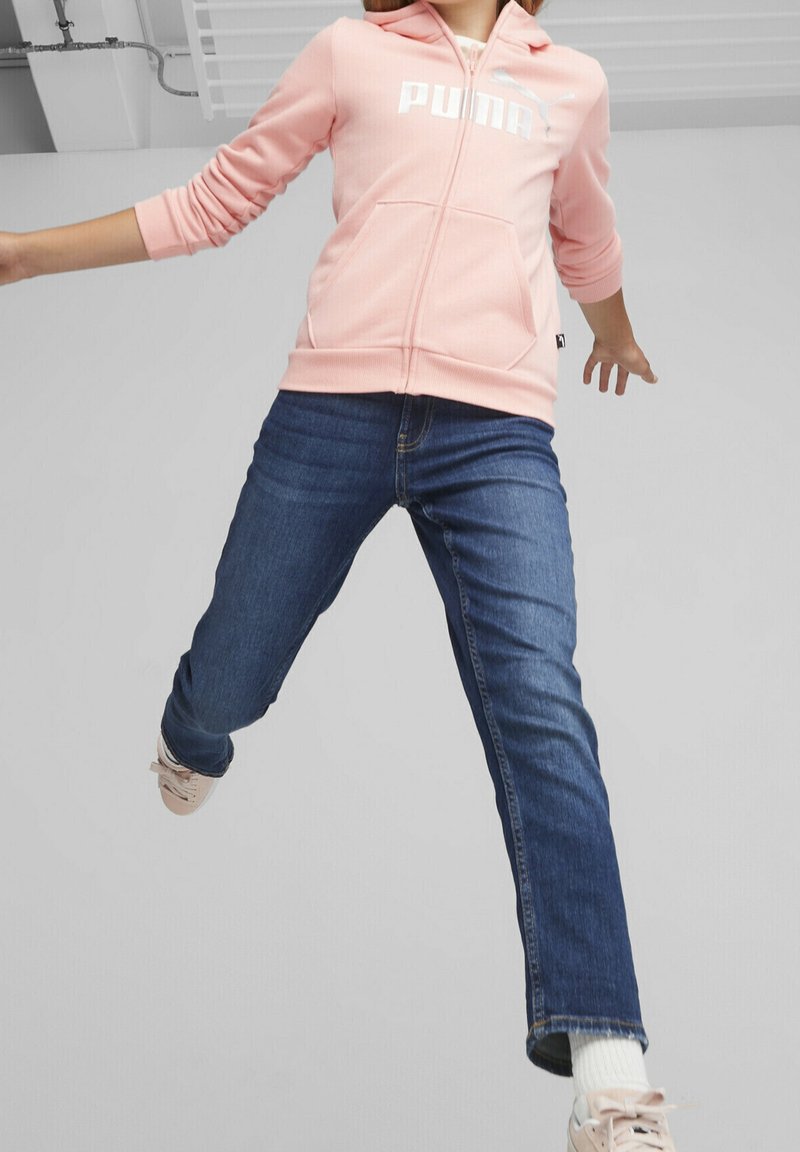 Sudadera de cremallera rosa clara con un logo blanco, combinada con jeans de denim azules y zapatillas rosas, capturada en plena acción contra un fondo neutro.