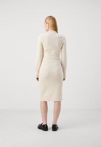 Abito lavorato a maglia aderente color crema con colletto alto e maniche lunghe, caratterizzato da una texture a coste e una cintura in vita. Sono indossate scarpe nere.