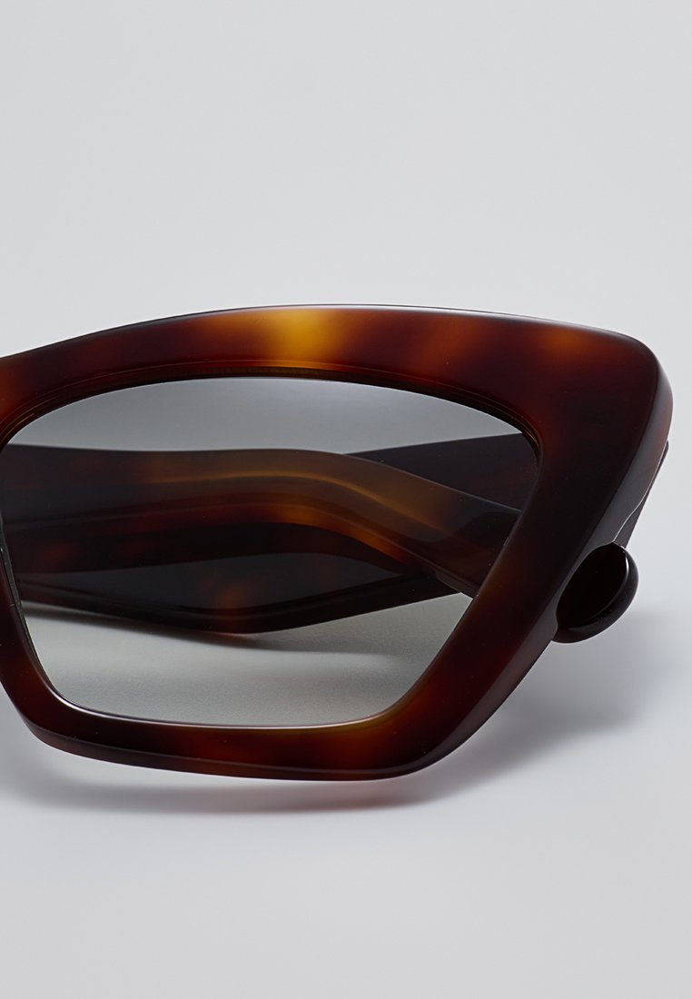 Gafas de sol carey con un marco angular y de gran tamaño. Las lentes tintadas oscuras presentan un diseño ondulado. Los acentos laterales incluyen un pequeño detalle circular.
