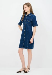 Denimmekko lyhyillä hihoilla, kauluskäänteellä, napitettavalla edessä, kahdella rintataskulla ja kireällä vyötäröllä. Tumma sininen väri ja sileä rakenne.