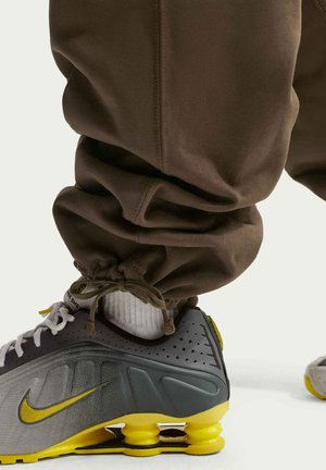 Graue und gelbe Nike-Sneaker mit strukturiertem Obermaterial, gepolsterter Sohle und sichtbarer Luftdämpfung. Braune Jogginghosen mit gerafften Knöcheln.