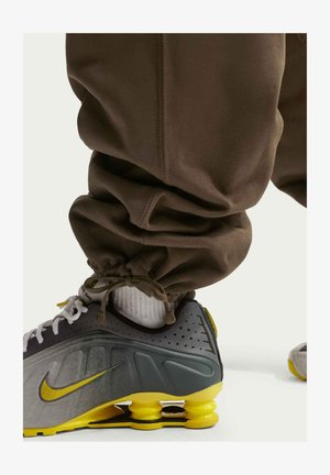 Graue und gelbe Nike-Sneaker mit strukturiertem Obermaterial, gepolsterter Sohle und sichtbarer Luftdämpfung. Braune Jogginghosen mit gerafften Knöcheln.