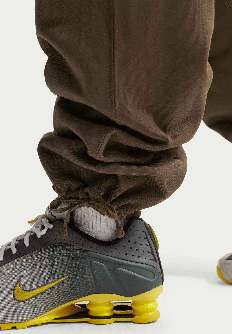 Scarpe da ginnastica Nike grigie e gialle con tomaia testurizzata, suola imbottita e ammortizzazione ad aria visibile. Pantaloni jogger marroni con caviglie raccolte.