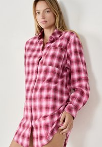 Różowa koszula w kratę z flaneli, z kołnierzykiem, kieszonką na piersi, zapinana na guziki, z długimi rękawami. Materiał wydaje się miękki i lekko teksturowany.