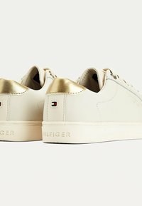 Białe skórzane sneakersy z złotymi akcentami na pięcie, charakteryzujące się teksturowanym tyłem, detalem z logo oraz płaską gumową podeszwą. Czysty design i sznurowanie.