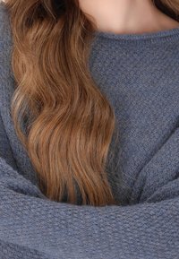 De longs cheveux bruns ondulés tombent sur un pull en tricot bleu foncé texturé, à coupe ample et manches larges.