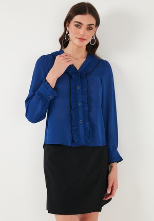 Blue blouse made of lightweight fabric featuring a ruffled front, button closures, and long sleeves with ruffled cuffs, paired with a black skirt.
