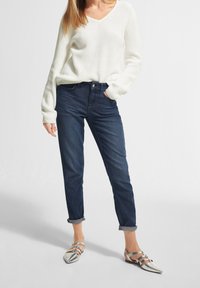 Biały sweter robiony na drutach z dekoltem w kształcie litery V, zestawiony z ciemnoniebieskimi, podwiniętymi dżinsami i srebrnymi płaskimi butami z krzyżującymi się paskami.