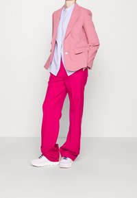 Blazer rosa claro sobre una camisa a rayas azul, combinado con pantalones rosas brillantes y zapatillas blancas. Presenta un corte holgado y una textura suave.