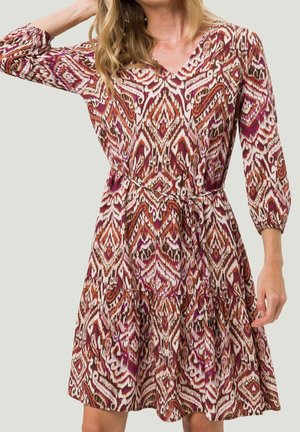 Femme portant une robe à manches longues avec un col en V, un motif géométrique rouge, marron et blanc, et une fine ceinture à la taille.