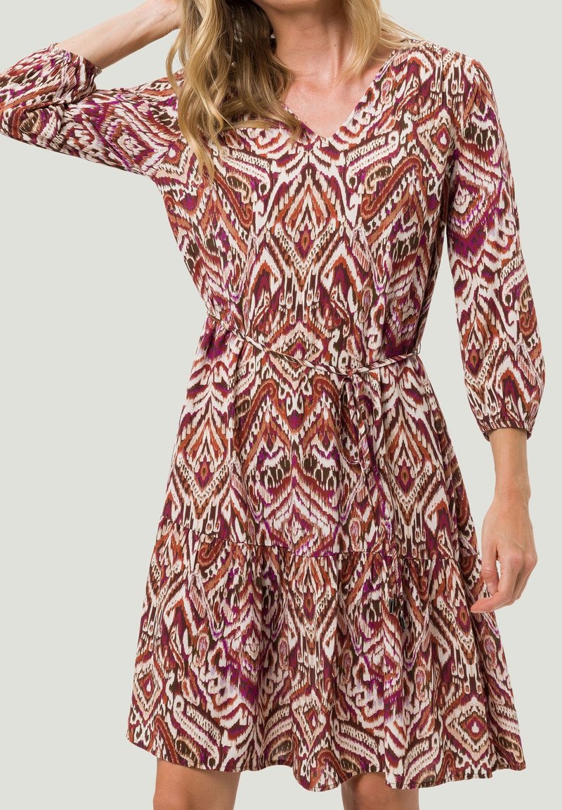 Femme portant une robe à manches longues avec un col en V, un motif géométrique rouge, marron et blanc, et une fine ceinture à la taille.