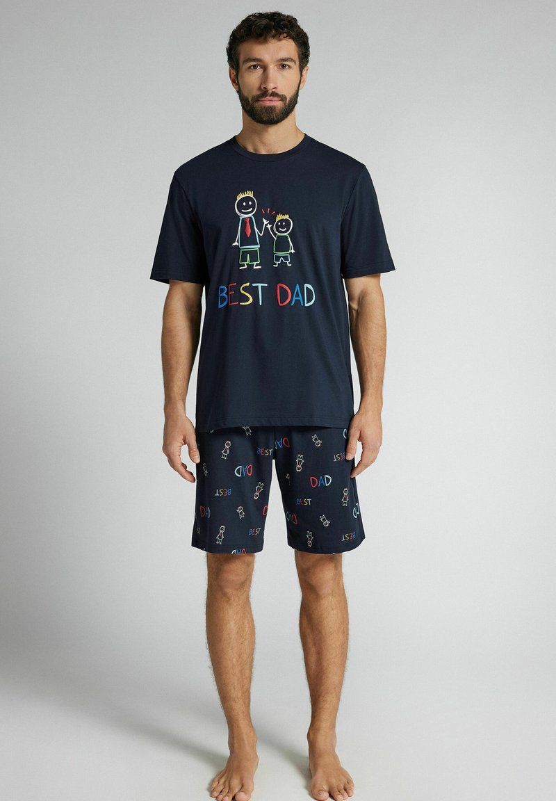 Mann trägt ein marineblaues Pyjama-Set mit dem Text "Bester Papa" und kindlichen Zeichnungen von zwei Strichmännchen, die sich auf dem Shirt und der Shorts an den Händen halten.