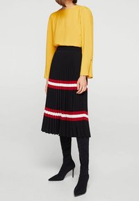 Blusa gialla a maniche lunghe con scollo rotondo abbinata a una gonna plissettata nera con righe orizzontali rosse e bianche, e stivali neri sopra il ginocchio.
