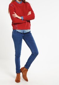 Pull rouge en maille avec détails en dentelle, chemise en denim en dessous, jean skinny bleu et bottines marron.