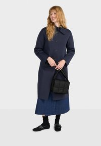 Merino-katso suuret napit ja kaulus, yhdistettynä mustaan tikattuun käsilaukkuun, farkkusortseihin, mustiin sukkiin ja korottomiin kenkiin, joissa on yksityiskohtia.