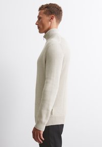 Beige gerippter Pullover mit hohem Kragen, ausgestattet mit einem Viertelreißverschluss und langen Ärmeln, kombiniert mit dunklen Hosen, seitlich betrachtet.