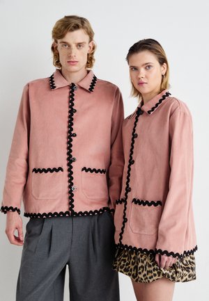 Roosa kangast jakk mustade nööpide ja taskute ääristustega, millel on kaks eesmist taskut; mudel on kombineerinud hallid püksid ja leopardimustriline seelik.