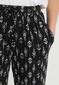 Zwarte broek met een wit geometrisch patroon, elastische tailleband met een trekkoord en zijzakken. Lichtgewicht stof en losse pasvorm.