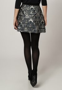 Femme portant une jupe à motifs floraux noirs et argentés, des collants noirs et des chaussures à talons hauts noires, se tenant devant un fond uni.