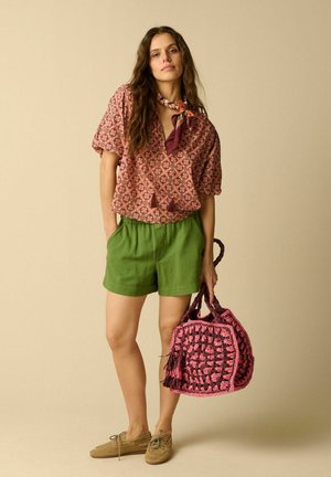 Femme portant un haut rouge à motifs, un short vert et une écharpe, tenant un sac crocheté rose et noir, debout devant un fond beige.